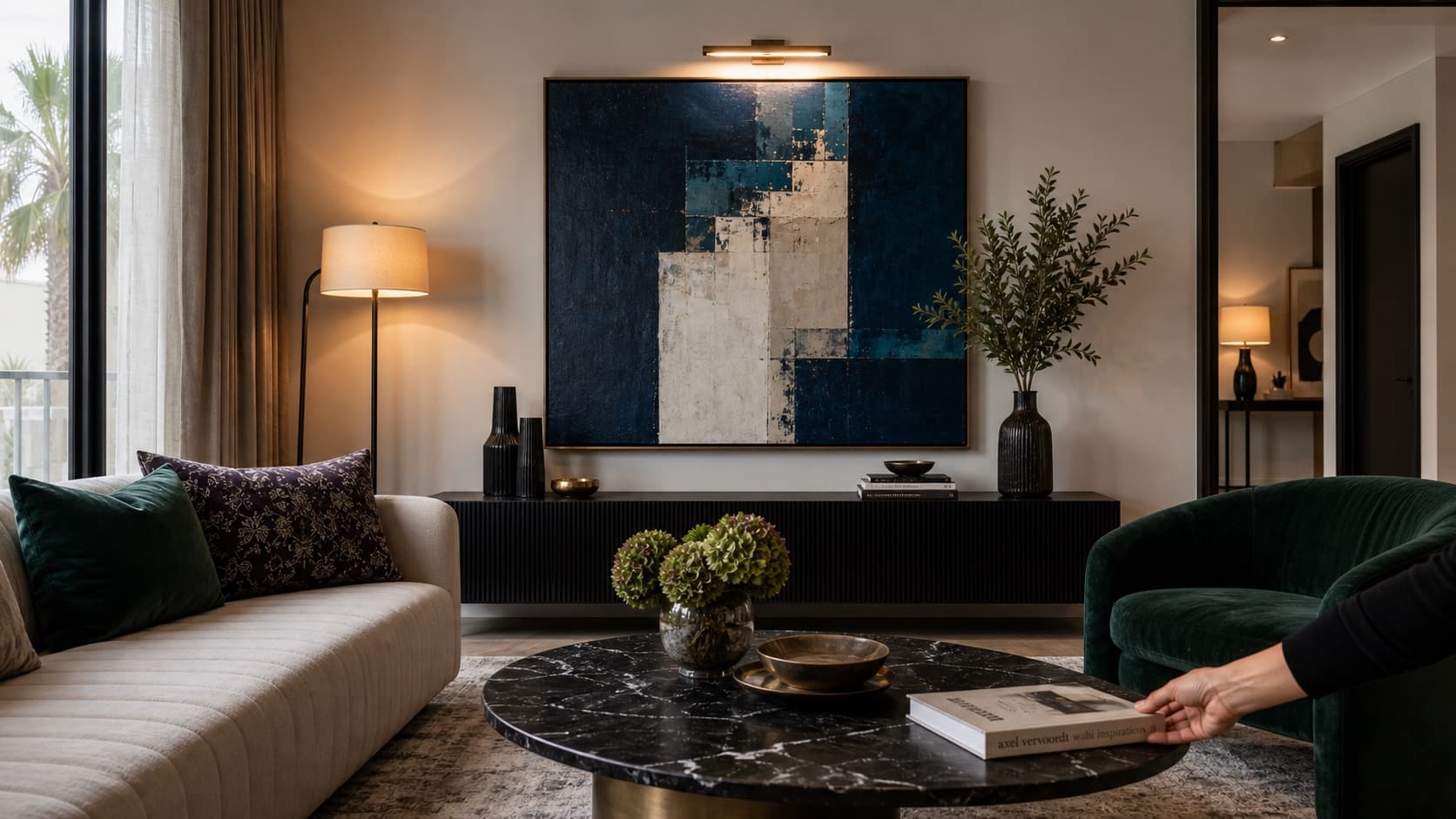 A refined living room with layered lighting, deep blue focal art, emerald accents, and a hand placing a sculptural bowl.
